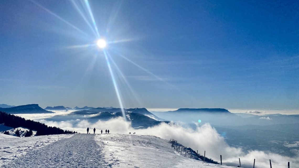 People walking along a snowy plateau in the French Alps under bright winter sun and clear blue sky.