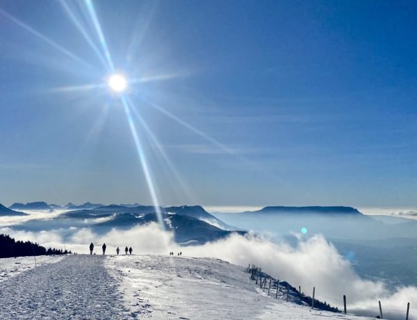 People walking along a snowy plateau in the French Alps under bright winter sun and clear blue sky.