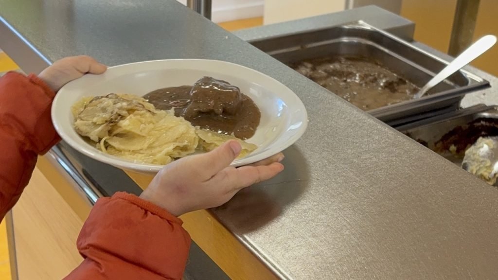 Child receiving a plated hot main course from the serving counter in a French school cafeteria.